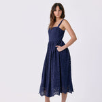 A woman modeling a navy blue lace midi dress with thin straps, side pockets, and a flared skirt, paired with white heeled sandals, against a plain light gray studio background.