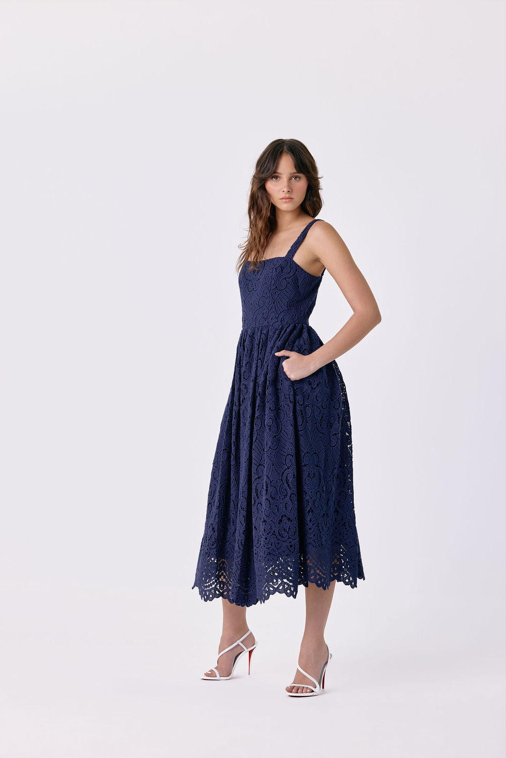 A woman modeling a navy blue lace midi dress with thin straps, side pockets, and a flared skirt, paired with white heeled sandals, against a plain light gray studio background.
