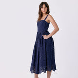 A woman modeling a navy blue lace midi dress with thin straps, side pockets, and a flared skirt, paired with white heeled sandals, against a plain light gray studio background.