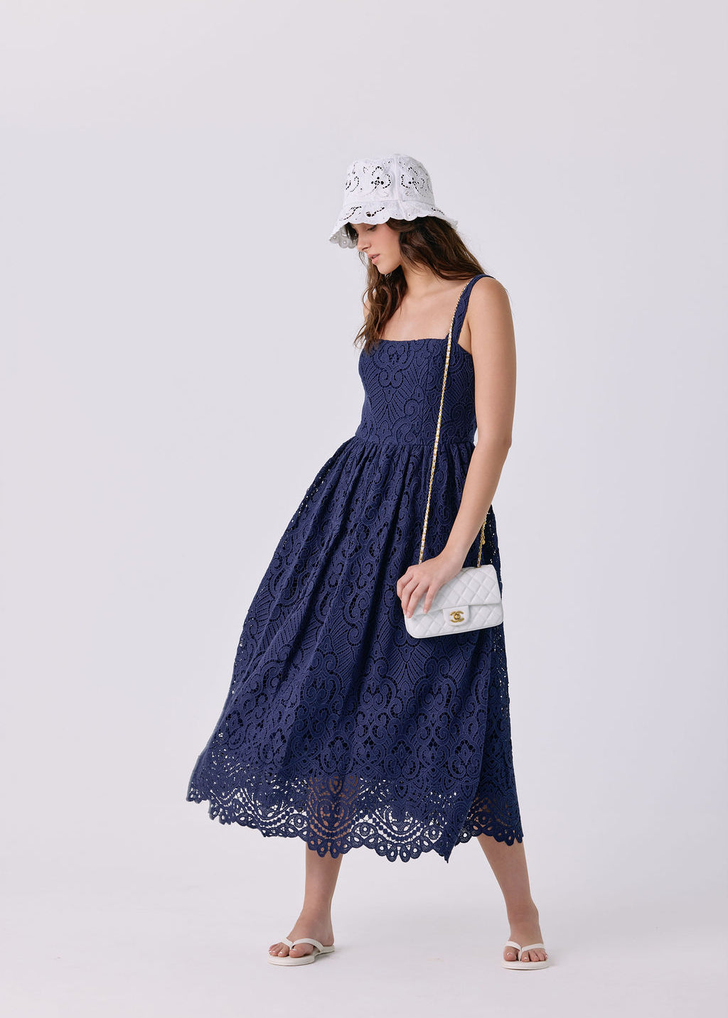 A woman modeling a navy blue lace midi dress with a gathered waist and thin straps, paired with a white bucket hat, a small white shoulder bag, and white slide sandals, against a light gray studio background.