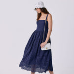 A woman modeling a navy blue lace midi dress with a gathered waist and thin straps, paired with a white bucket hat, a small white shoulder bag, and white slide sandals, against a light gray studio background.