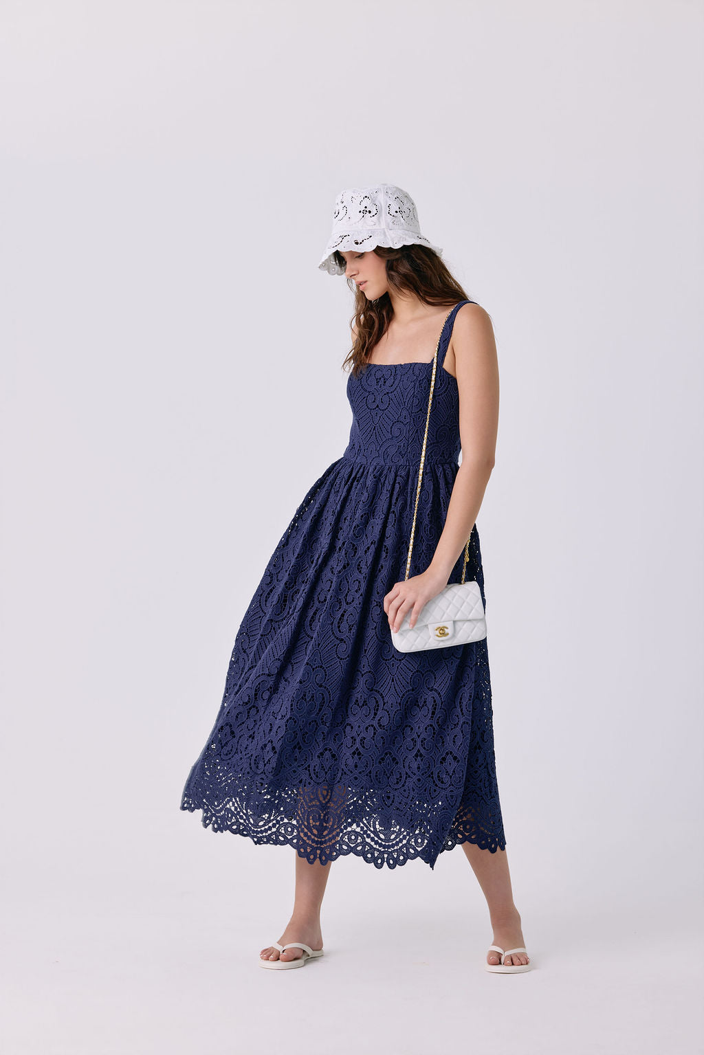 A woman modeling a navy blue lace midi dress with a gathered waist and thin straps, paired with a white bucket hat, a small white shoulder bag, and white slide sandals, against a light gray studio background.