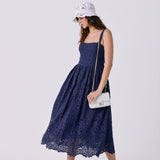 A woman modeling a navy blue lace midi dress with a gathered waist and thin straps, paired with a white bucket hat, a small white shoulder bag, and white slide sandals, against a light gray studio background.