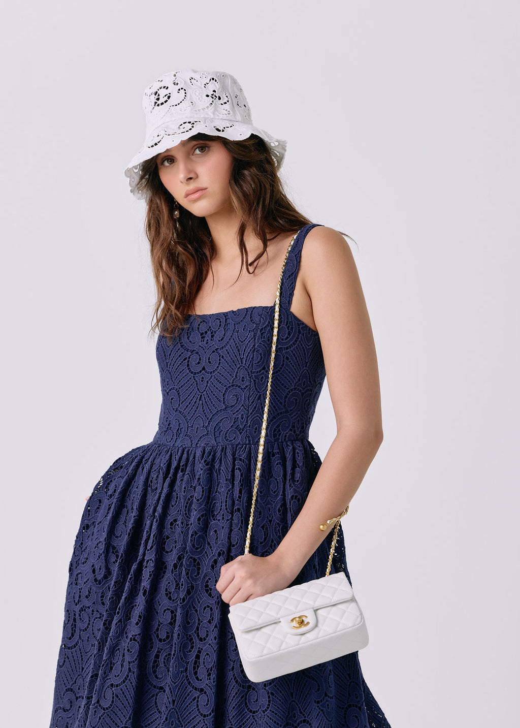 A woman wearing a navy blue lace dress, a white bucket hat with black floral embroidery, and holding a white quilted shoulder bag with a gold chain strap.