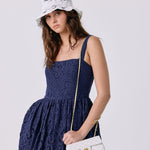 A woman wearing a navy blue lace dress, a white bucket hat with black floral embroidery, and holding a white quilted shoulder bag with a gold chain strap.