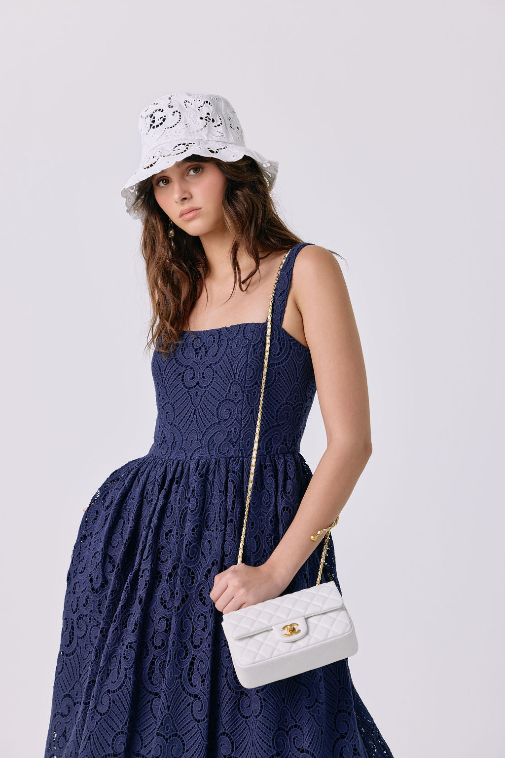A woman wearing a navy blue lace dress, a white bucket hat with black floral embroidery, and holding a white quilted shoulder bag with a gold chain strap.