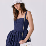 A woman wearing a navy blue lace dress, a white bucket hat with black floral embroidery, and holding a white quilted shoulder bag with a gold chain strap.