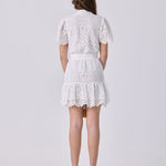 A woman viewed from the back wearing a white lace short dress with short puff sleeves and a ruffled hem, paired with red high heels, against a light gray studio background.