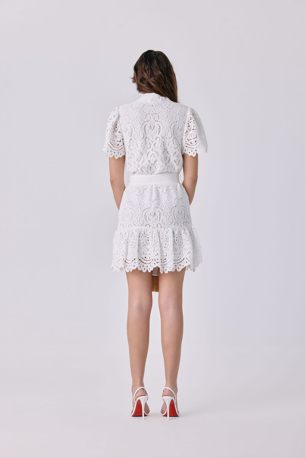 A woman viewed from the back wearing a white lace short dress with short puff sleeves and a ruffled hem, paired with red high heels, against a light gray studio background.