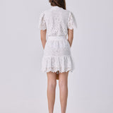 A woman viewed from the back wearing a white lace short dress with short puff sleeves and a ruffled hem, paired with red high heels, against a light gray studio background.