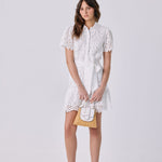A woman modeling a white short-sleeve lace dress with a collar and matching belt, holding a small white and gold handbag, against a light gray studio background.