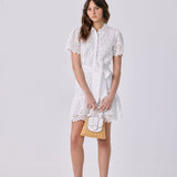 A woman modeling a white short-sleeve lace dress with a collar and matching belt, holding a small white and gold handbag, against a light gray studio background.