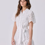 A woman modeling a white short-sleeve lace mini dress with a tie waist, standing against a light gray studio background and holding a small structured handbag.