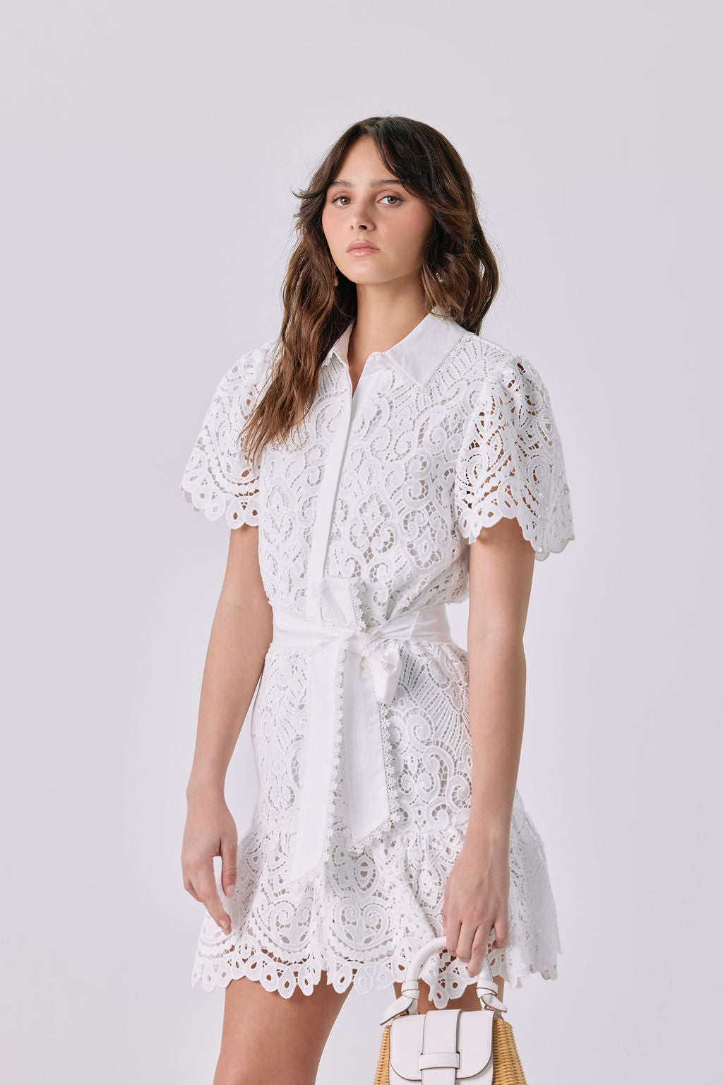 A woman modeling a white short-sleeve lace mini dress with a tie waist, standing against a light gray studio background and holding a small structured handbag.