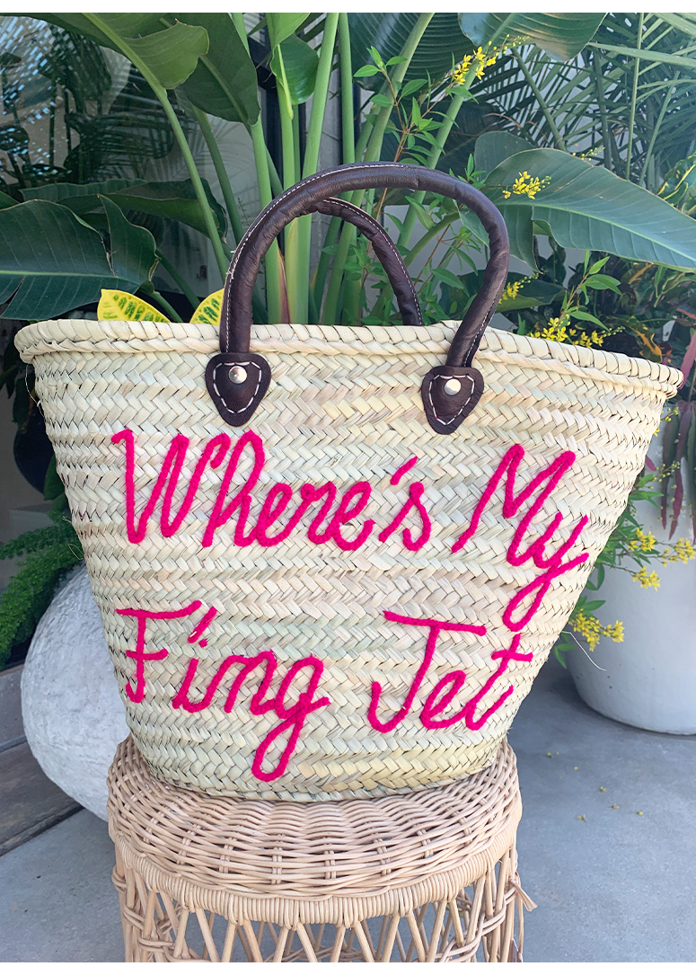 A straw tote bag with brown handles and the text 'Where's My F'ing Jet' in pink yarn on the side, displayed on a wicker surface with greenery in the background.